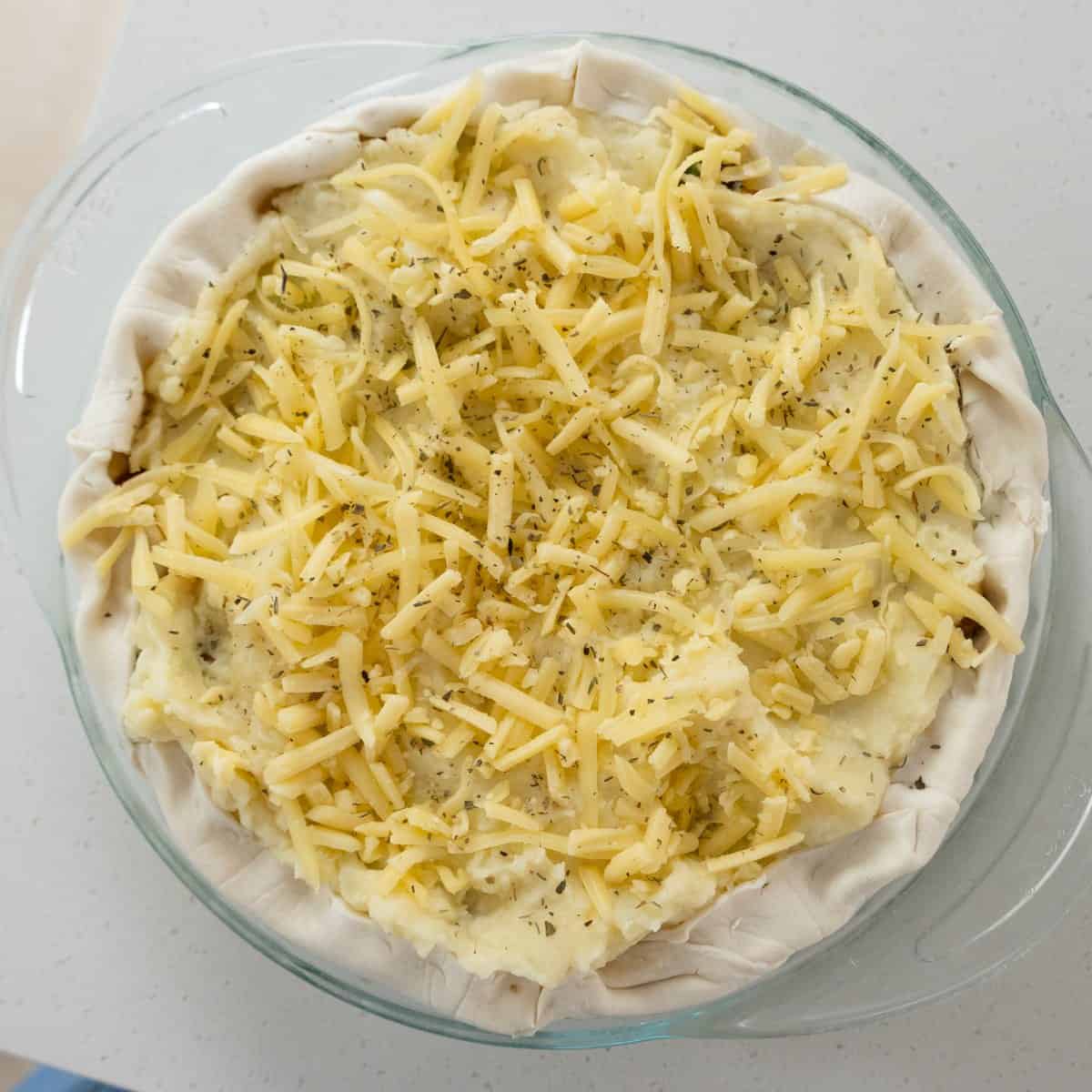 Shepherd's Pie Unbaked pie in a glass dish with a pastry crust, topped with mashed potatoes, shredded cheese, and black pepper.