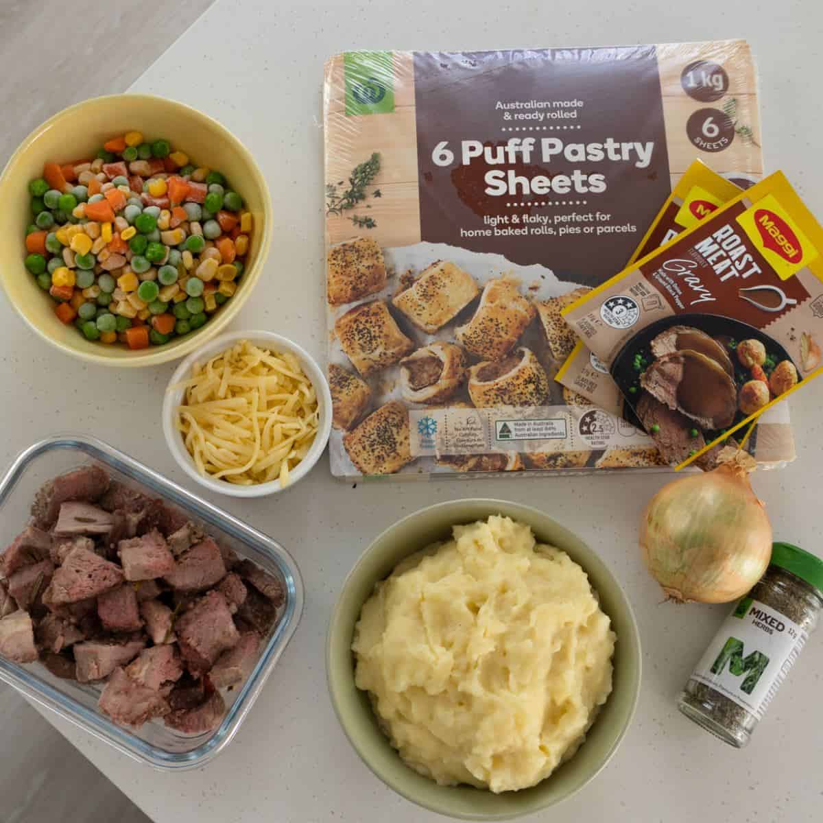Shepherd's Pie Ingredients on a counter include mixed vegetables, shredded cheese, cooked meat, mashed potatoes, puff pastry sheets, gravy mix, an onion, and mixed herbs.