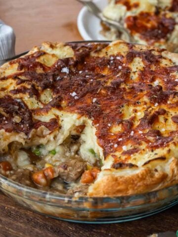 A glass pie dish filled with a savory pie topped with browned mashed potatoes, showing a portion removed to reveal meat, peas, and carrots inside.