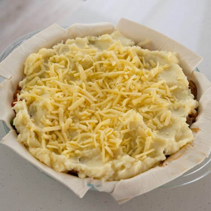 Shepherd's Pie Unbaked pie topped with mashed potatoes and shredded cheese, placed in a parchment-lined glass dish on a white countertop.