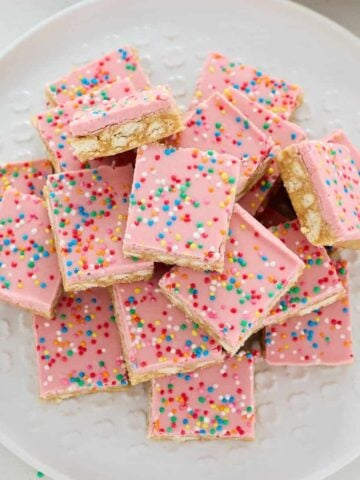 A plate of pink frosted dessert bars topped with colorful round sprinkles, arranged in a pile on a white plate.
