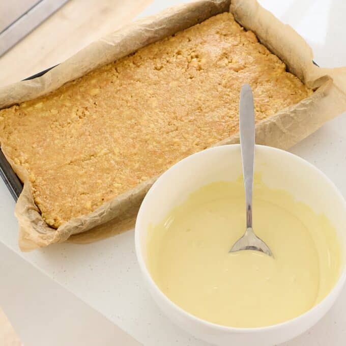 Sprinkle Biscuit Slice A rectangular baked oat base in a parchment-lined tray next to a white bowl of pale yellow icing with a spoon.