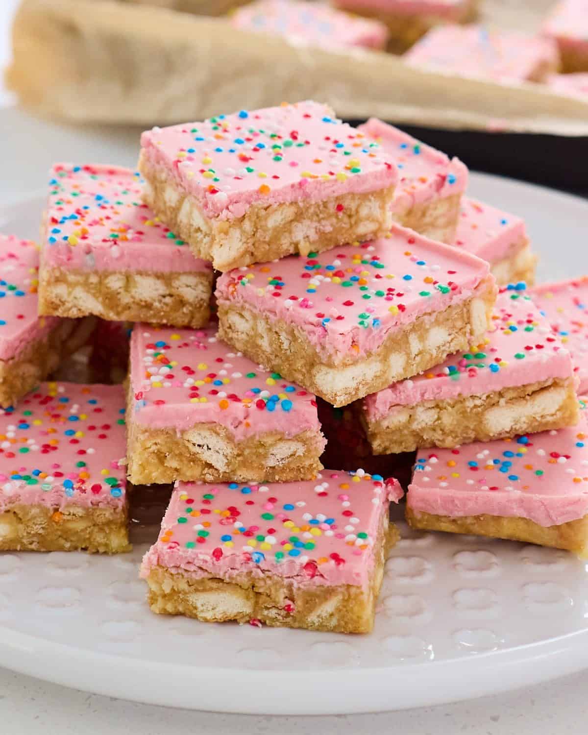 A stack of pink-frosted dessert bars with colorful sprinkles is arranged on a white plate.