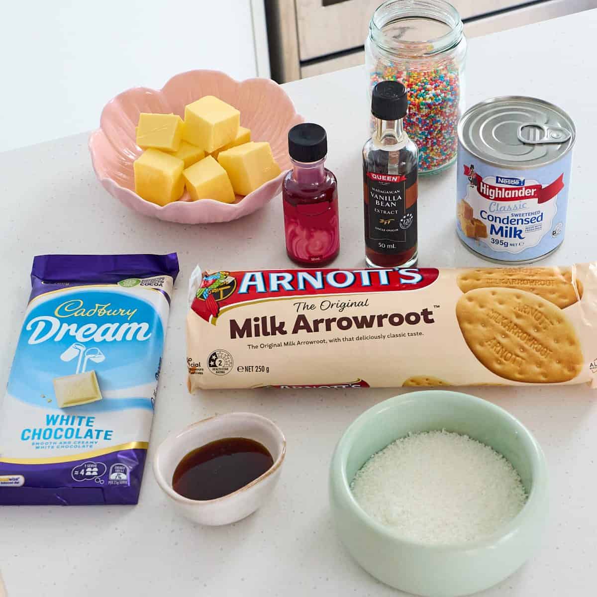 Assorted ingredients for baking, including white chocolate, milk arrowroot biscuits, butter, condensed milk, desiccated coconut, vanilla, food coloring, and rainbow sprinkles on a countertop.