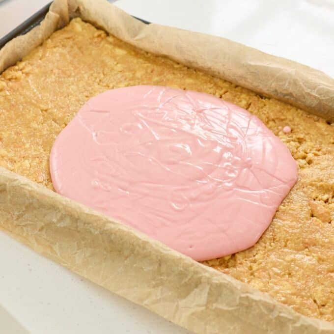 Sprinkle Biscuit Slice A rectangular tray of baked biscuit base lined with parchment paper, topped with a large pool of pink icing in the center, ready to be spread.