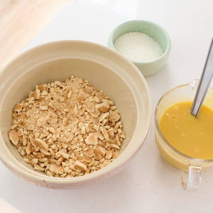Sprinkle Biscuit Slice A bowl of crushed biscuits, a small bowl of sugar, and a measuring cup with melted butter and a spoon, arranged on a white countertop.