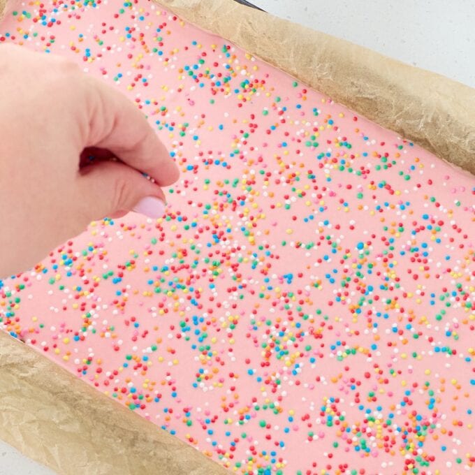 Sprinkle Biscuit Slice A hand sprinkles colorful round sprinkles onto a sheet of pink frosted dessert in a parchment-lined baking tray.