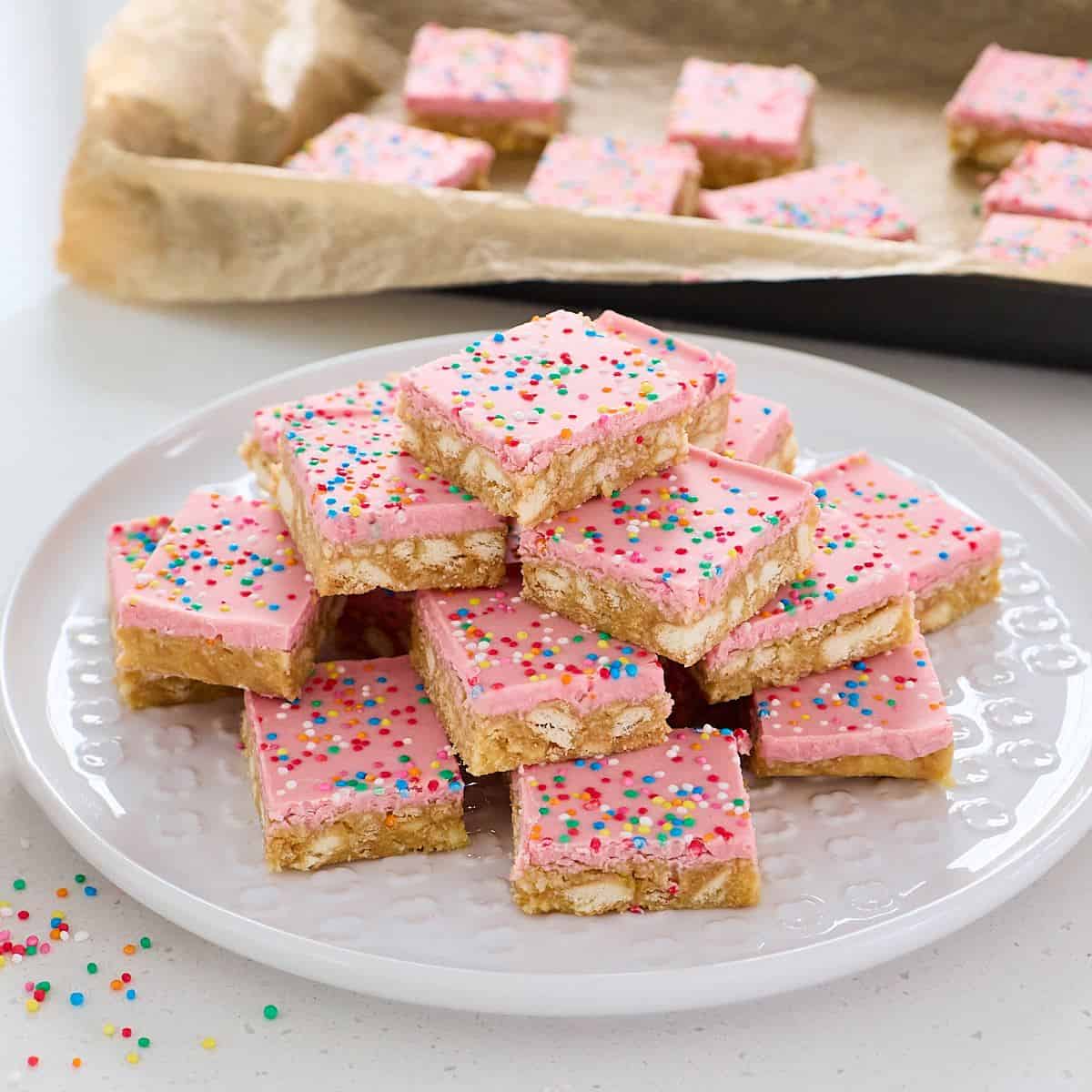Sprinkle Biscuit Slice A plate of frosted dessert bars with pink icing and colorful sprinkles is stacked in the foreground, with more bars in a pan lined with parchment paper in the background.