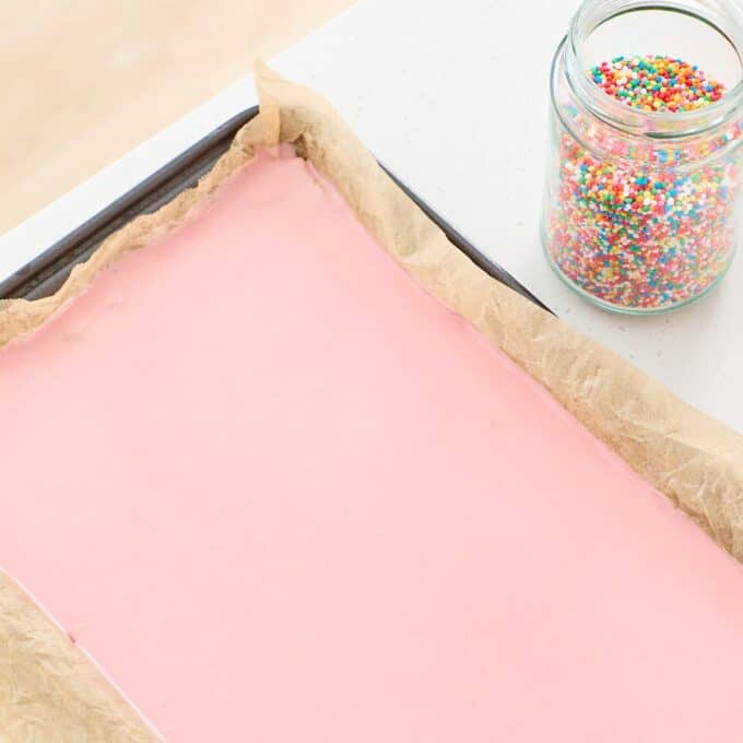 Sprinkle Biscuit Slice A baking tray lined with parchment paper holds a layer of smooth pink batter; a jar of colorful round sprinkles sits nearby on the counter.
