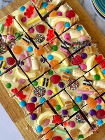A tray of white chocolate slices topped with assorted colorful candies, cut into squares and arranged on a wooden board next to stacked plates.
