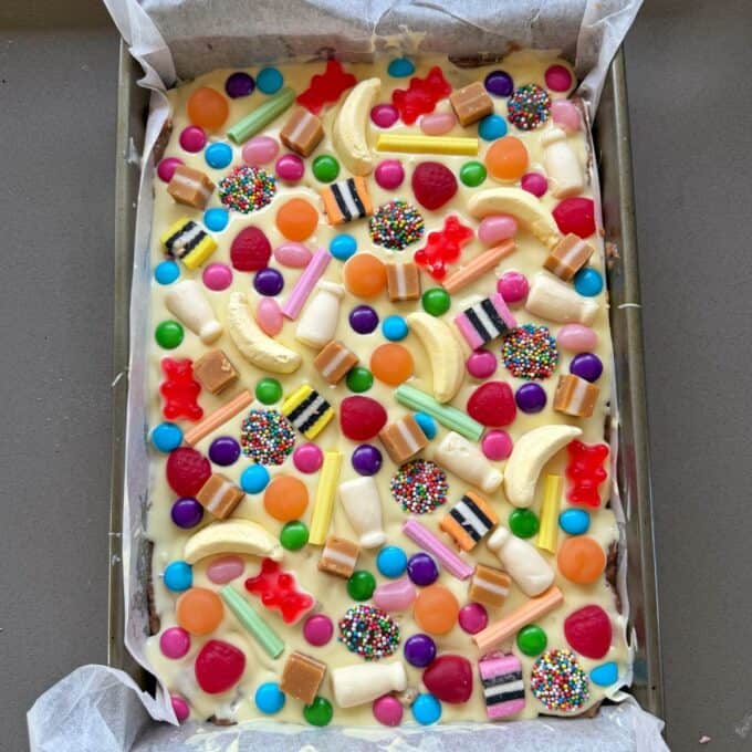 XL Pick and Mix Lolly Slice A rectangular baking tray filled with white chocolate slice, topped with colorful candies, chocolates, jelly sweets, and licorice pieces on parchment paper.