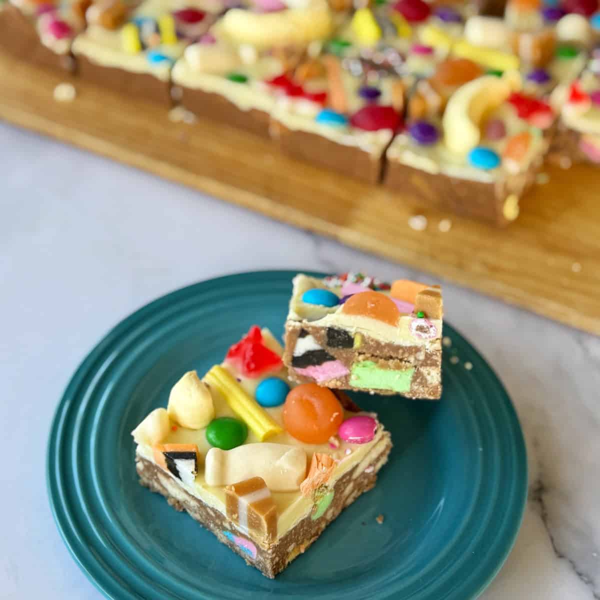 XL Pick and Mix Lolly Slice Two colorful slices of candy-topped dessert bars rest on a teal plate, with more bars arranged on a wooden board in the background.