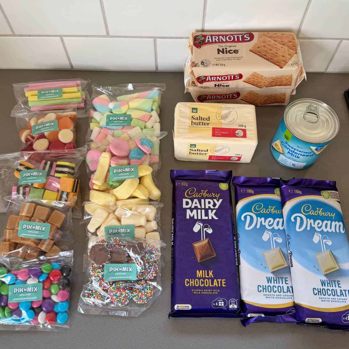 XL Pick and Mix Lolly Slice Assorted confectionery, biscuits, two blocks of butter, a can of condensed milk, three milk chocolate bars, and three white chocolate bars arranged on a kitchen counter.