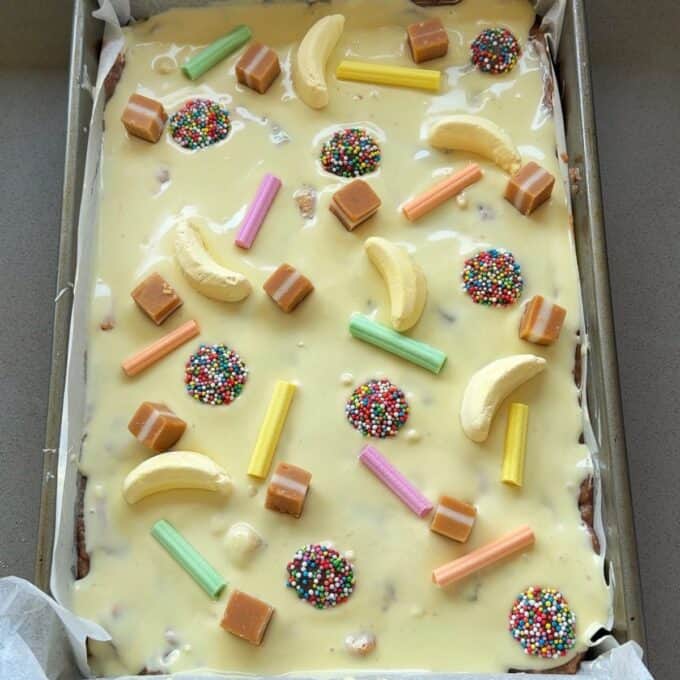 XL Pick and Mix Lolly Slice A tray of slice covered in pale yellow icing and topped with banana candies, caramel cubes, rainbow sprinkle discs, and colorful candy sticks.