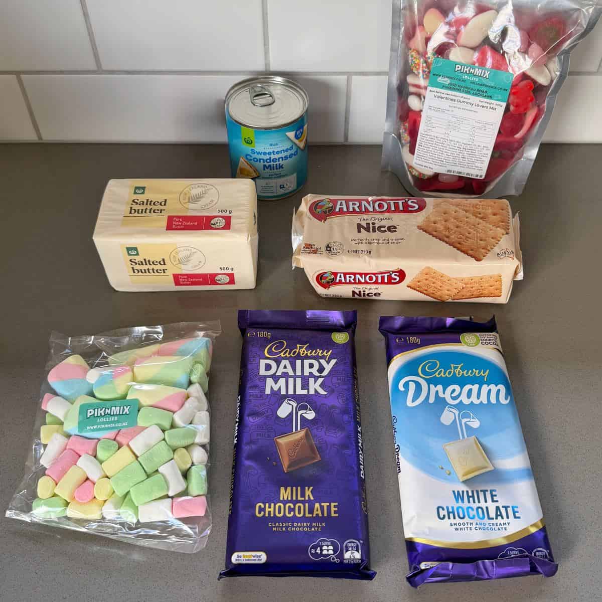 Assorted ingredients on a countertop, including butter, sweetened condensed milk, Nice biscuits, strawberry candy, assorted marshmallows, milk chocolate, and white chocolate.