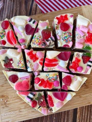 Heart-shaped dessert topped with white chocolate, colorful candies, gummies, and sprinkles, cut into 12 pieces on a wooden cutting board.