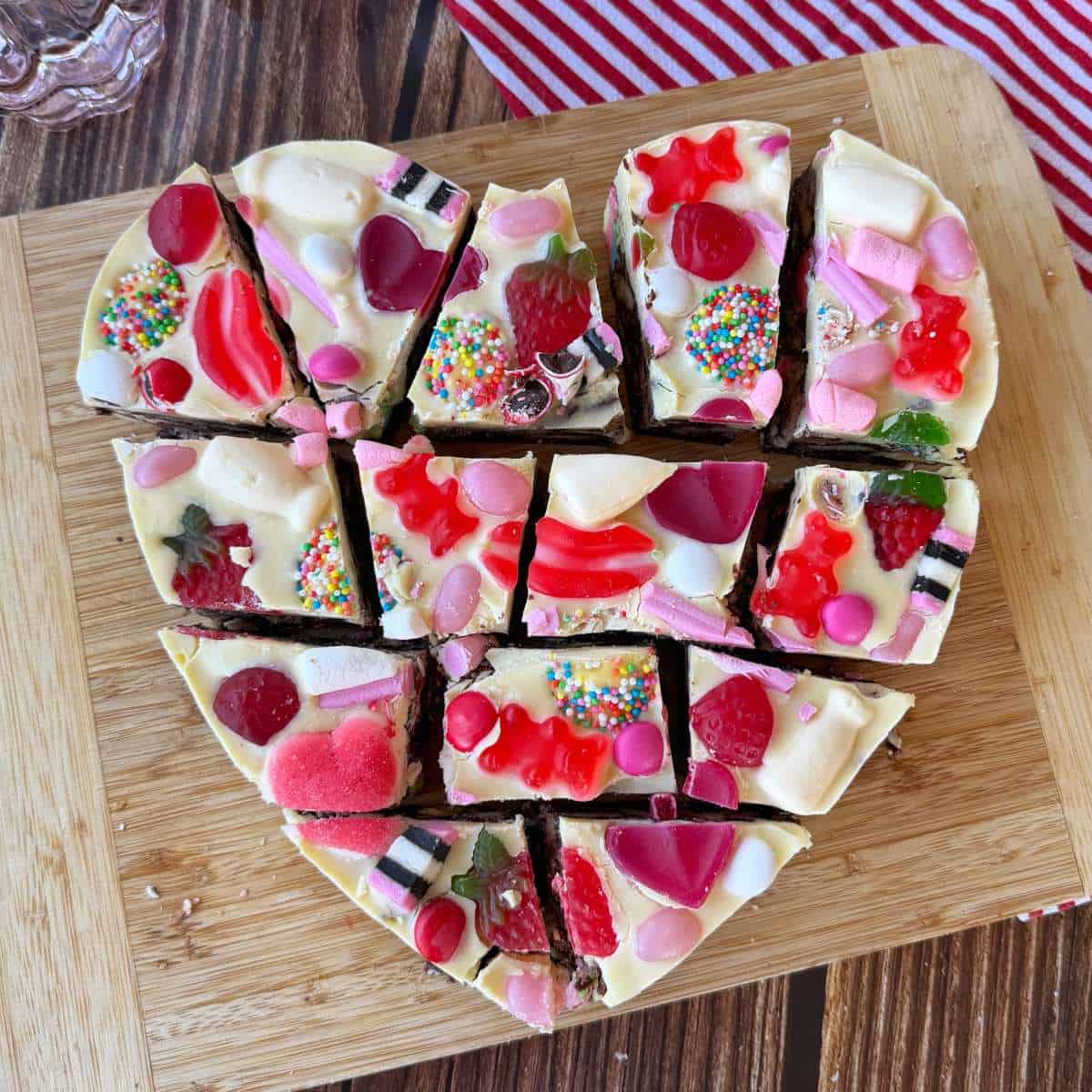 Love Heart Lolly Slice Heart-shaped dessert topped with white chocolate, colorful candies, gummies, and sprinkles, cut into 12 pieces on a wooden cutting board.