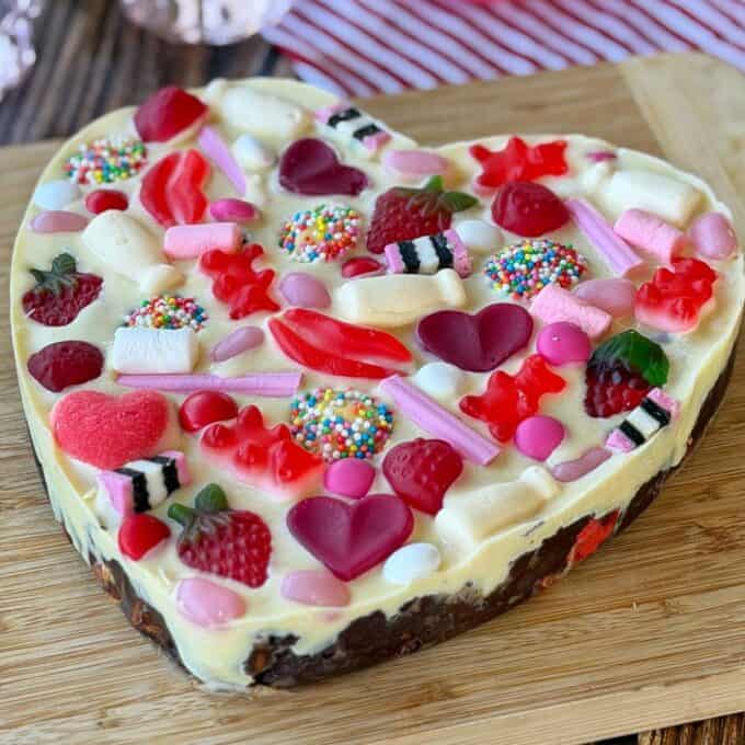 Love Heart Lolly Slice Heart-shaped dessert topped with assorted colorful candies, gummies, sprinkles, and chocolates, placed on a wooden board.