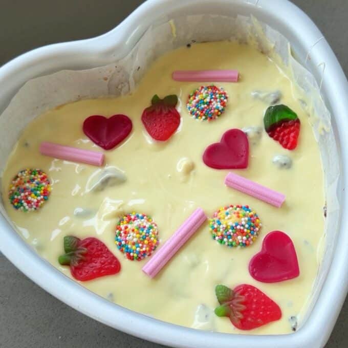 Love Heart Lolly Slice Heart-shaped cake pan filled with cake batter, topped with gummy hearts, gummy strawberries, pink candy sticks, and round chocolate candies with rainbow sprinkles.