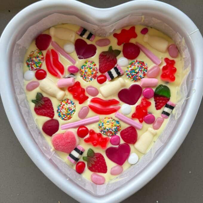 Love Heart Lolly Slice A heart-shaped cake tin filled with cake batter, topped with assorted colorful candies, gummies, and sprinkles, ready to be baked.
