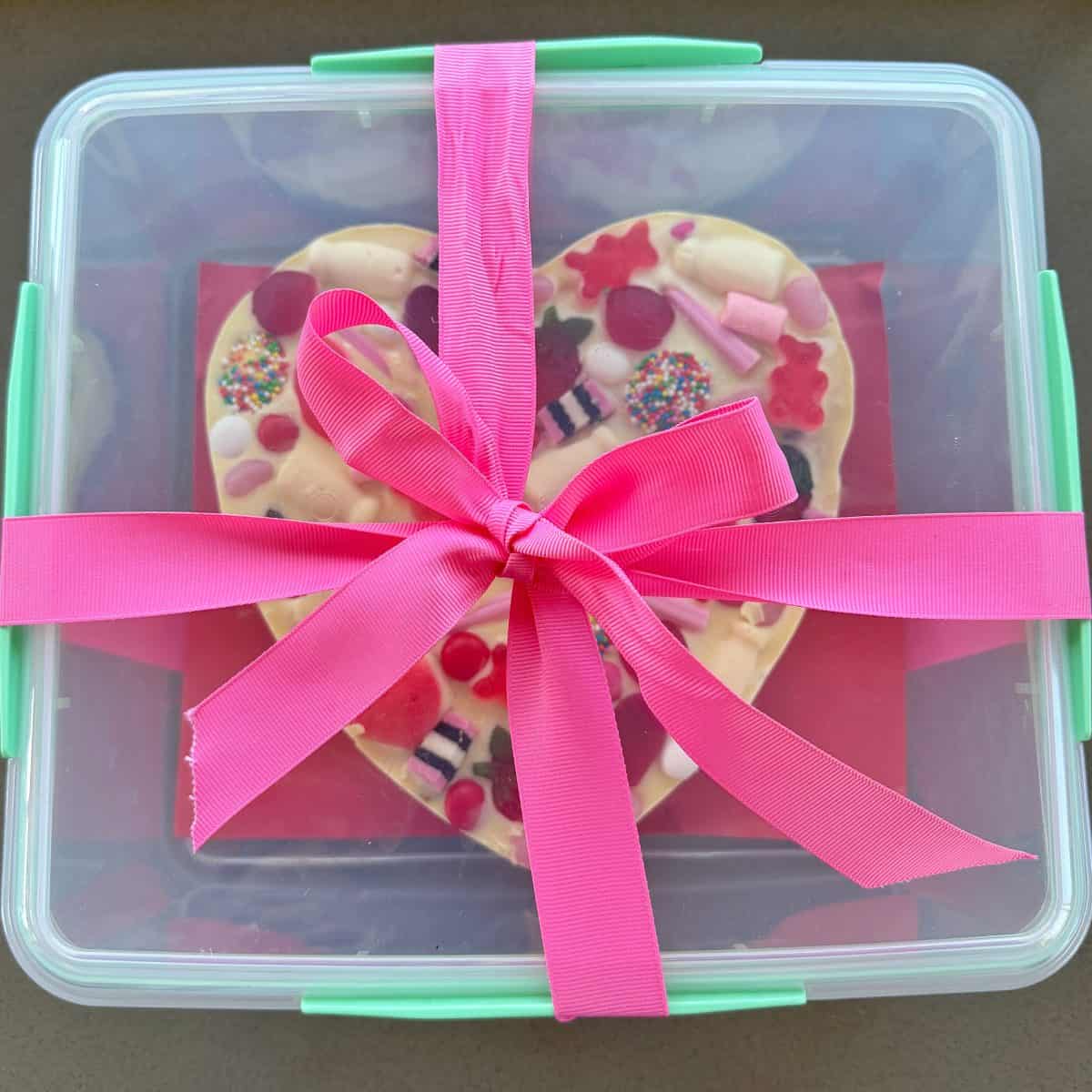 A clear plastic container with green clasps holds a heart-shaped cookie decorated with colorful candies, tied with a bright pink ribbon.