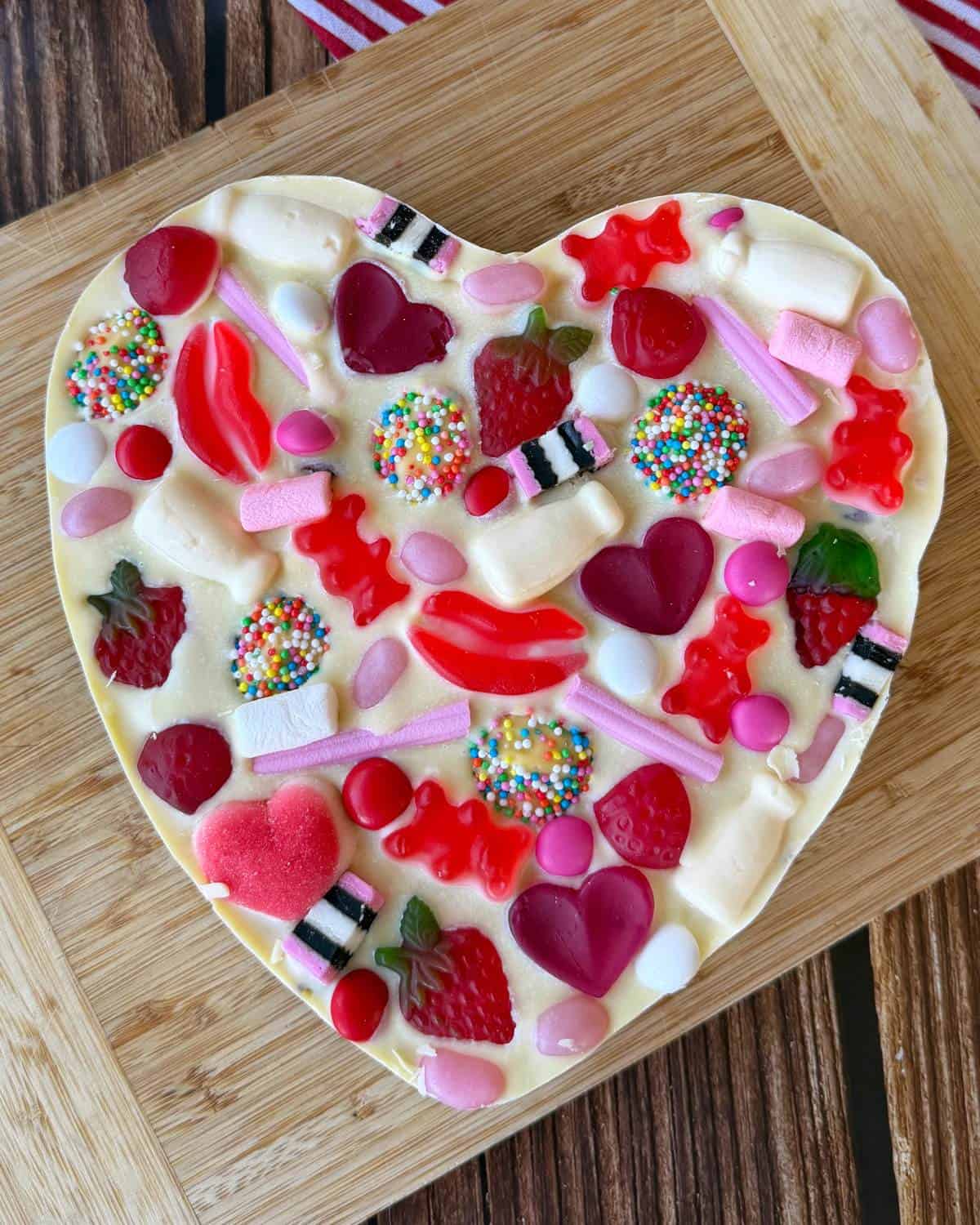 A heart-shaped white chocolate slab topped with various colorful candies, gummies, and sprinkles, placed on a wooden cutting board.