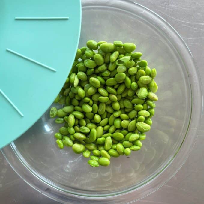 Zesty Tuna, Avocado and Edamame Salad A glass bowl containing green edamame beans is partially covered by a blue lid, placed on a metallic surface.