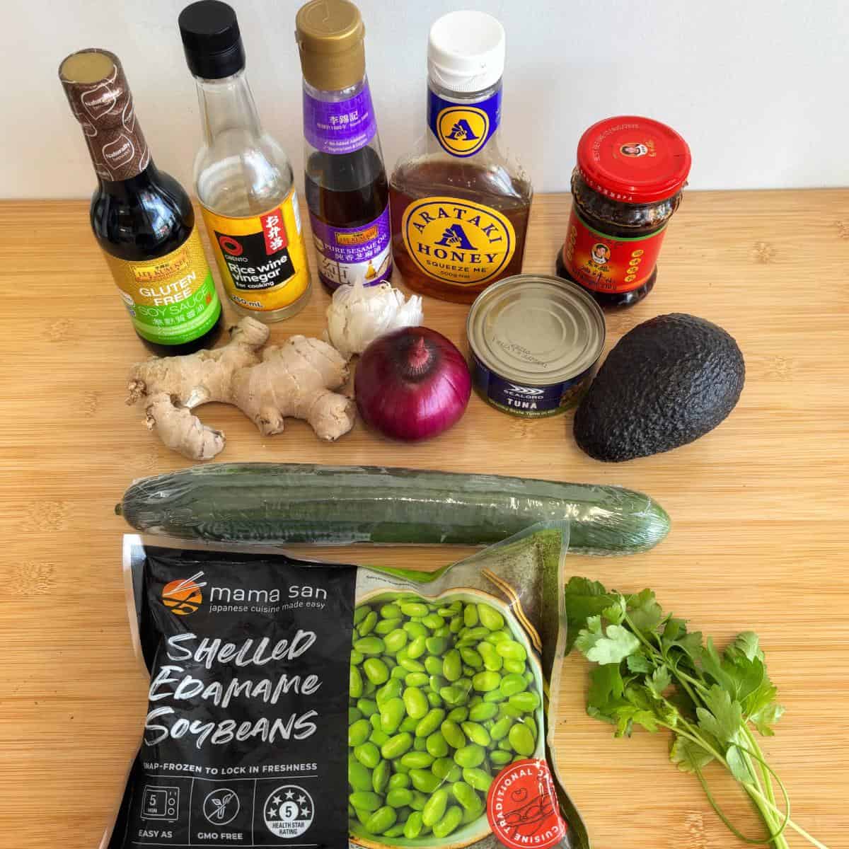 Zesty Tuna, Avocado and Edamame Salad A selection of ingredients on a wooden surface, including sauces, ginger, red onion, avocado, cucumber, cilantro, edamame, honey, tuna, and chili garlic sauce.