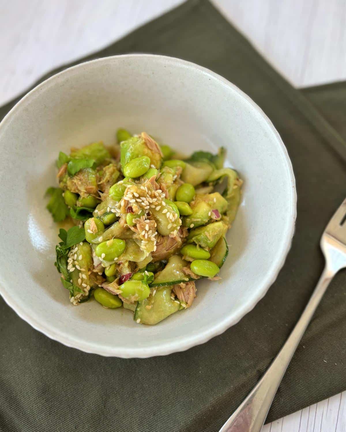 A bowl of salad with edamame, cucumber, shredded chicken, avocado, and sesame seeds, placed on a green napkin beside a fork.
