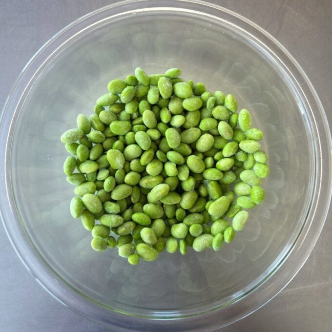 Zesty Tuna, Avocado and Edamame Salad A clear glass bowl filled with frozen green edamame beans sits on a gray surface.