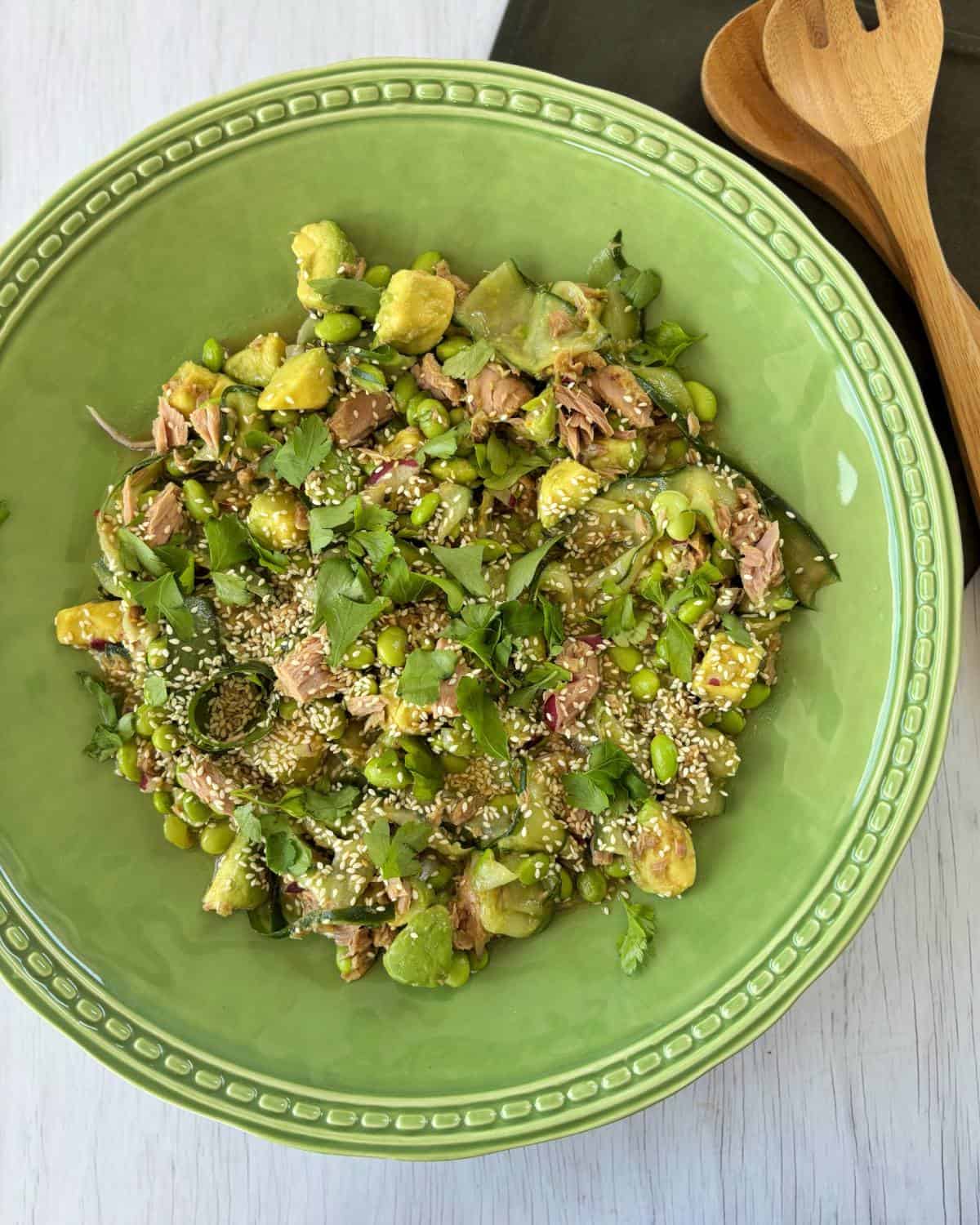 Zesty Tuna, Avocado and Edamame Salad Green ceramic bowl filled with a salad containing avocado, edamame, tuna, sesame seeds, chopped herbs, and mixed greens, placed on a light wooden surface next to wooden utensils.