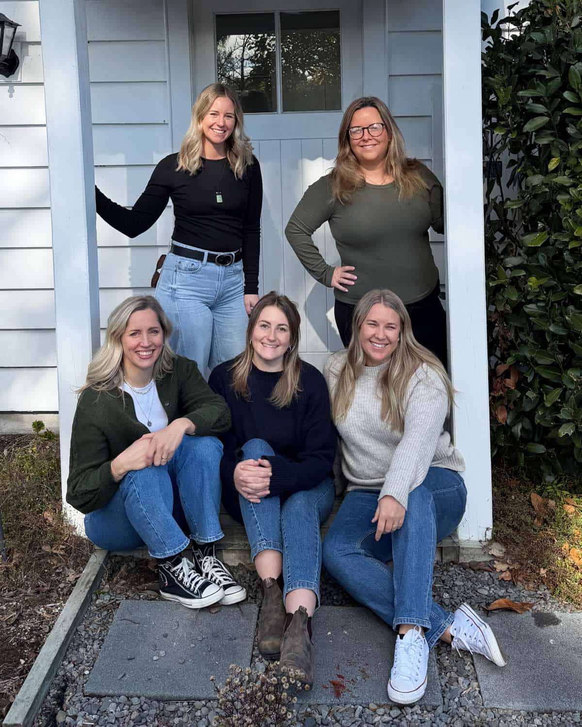 Five women pose together outside a house, with two standing in the doorway and three sitting on the front steps. All are smiling and dressed in casual sweaters and jeans.