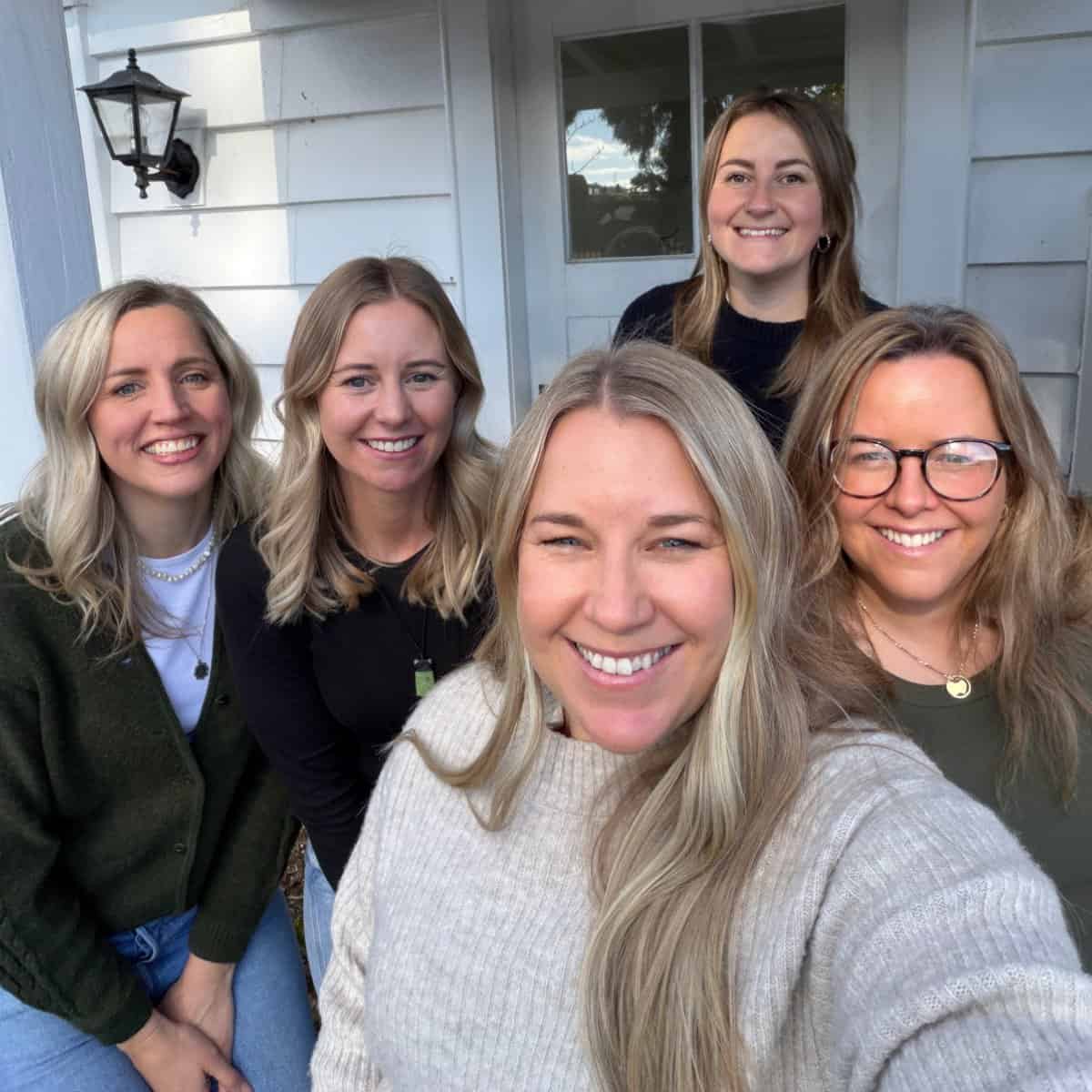 Five women pose and smile for a group photo outside a white house with horizontal siding and a lantern mounted on the wall.