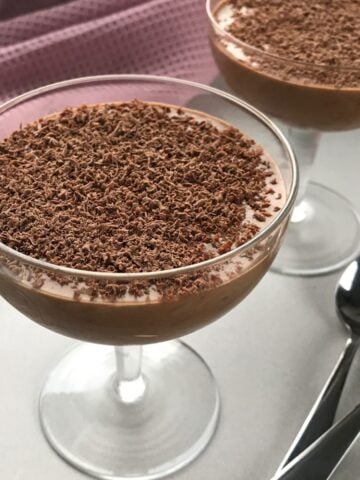 Two glass dessert cups filled with rich chocolate mousse, topped with grated chocolate, with two metal spoons beside them on a white surface.