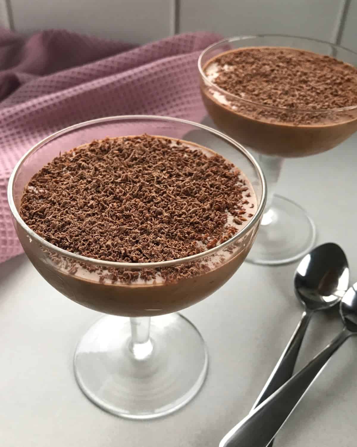 Simple Chocolate Mousse Two glass dessert cups filled with chocolate mousse, topped with grated chocolate, placed on a light surface beside two metal spoons and a purple textured cloth in the background.