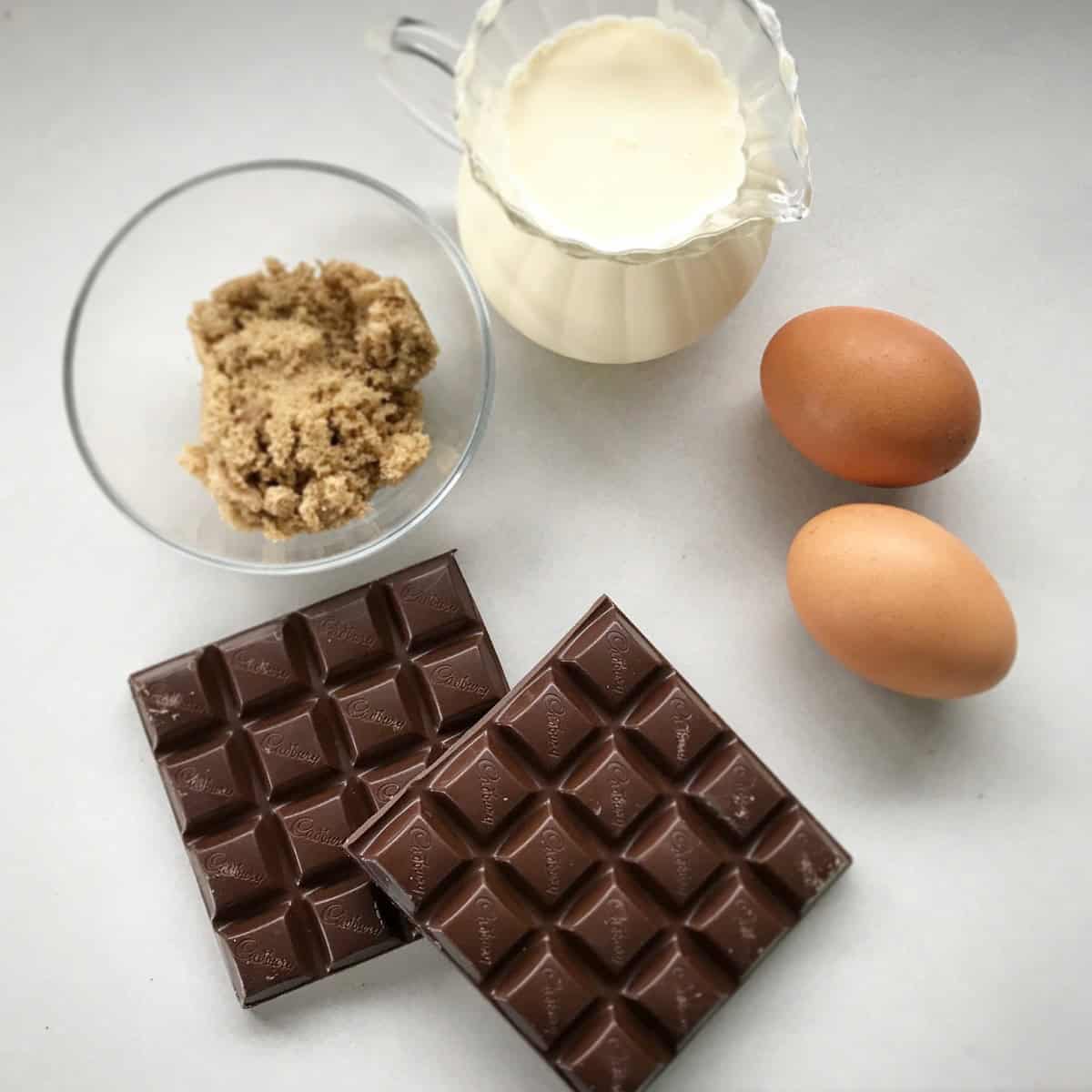 Simple Chocolate Mousse A glass jug of cream, two eggs, a bowl of brown sugar, and two bars of chocolate arranged on a light surface.