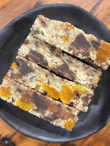 Three rectangular dessert bars with visible chunks of dried apricot and chocolate are stacked on a black plate, placed on a wooden surface.