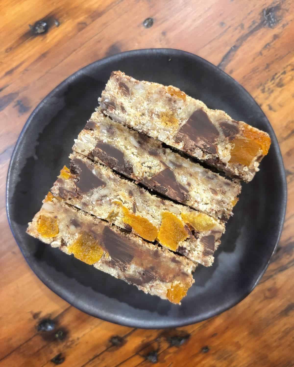 Gluten Free Bumper Bars Three rectangular slices of oat bars with visible chocolate chunks and dried apricots are arranged on a black plate atop a wooden table.