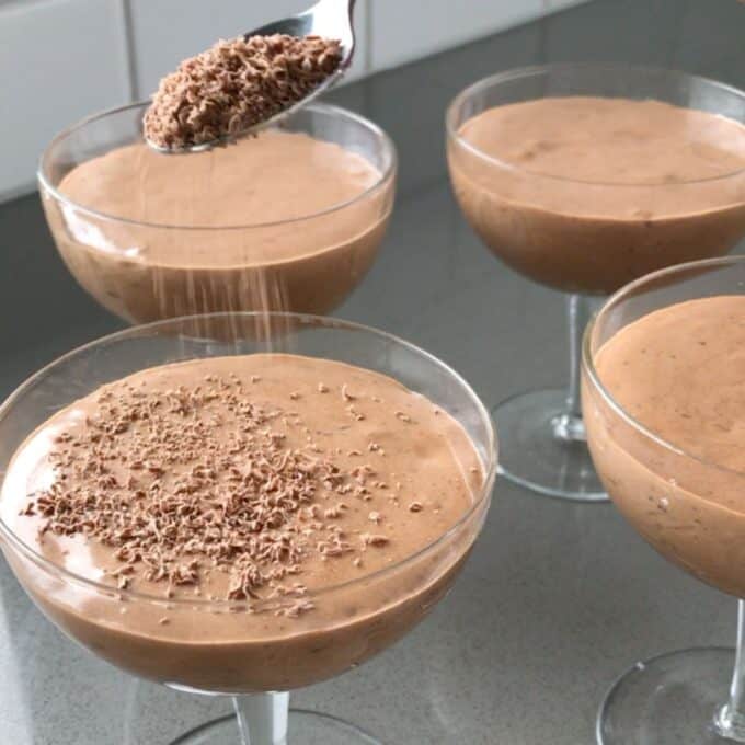 Simple Chocolate Mousse Four glass bowls filled with chocolate mousse; a spoon is sprinkling grated chocolate onto one of the servings on a kitchen countertop.