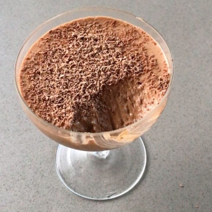 Simple Chocolate Mousse A glass dish of chocolate mousse topped with grated chocolate, with a spoonful removed, on a gray surface.