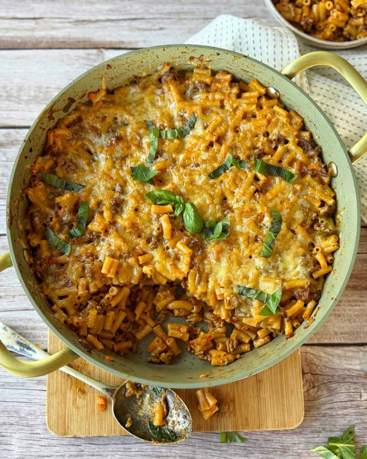 Cheesy Taco Mince and Macaroni Bake A large pan of baked macaroni and cheese with a golden-brown cheesy top, garnished with fresh basil, sits on a wooden board with a serving spoon and a small bowl nearby.