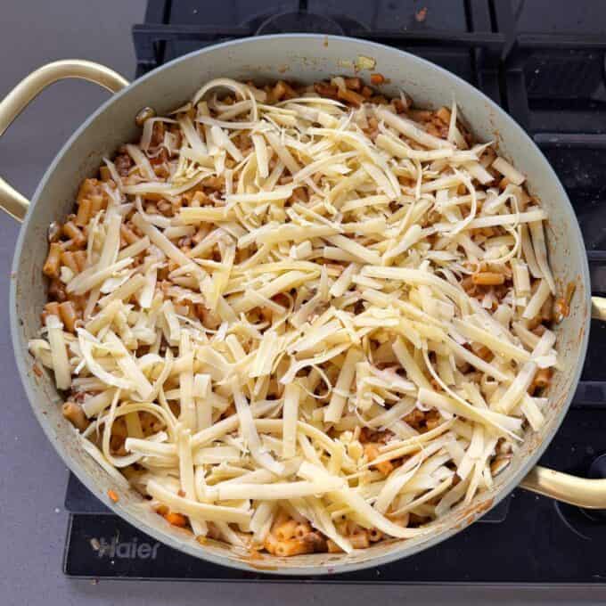 Cheesy Taco Mince and Macaroni Bake A large round pan on a stove contains pasta mixed with sauce, topped with a generous layer of shredded cheese, ready to be baked or melted.