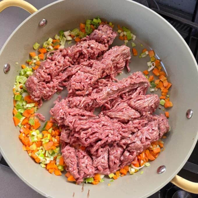 Cheesy Taco Mince and Macaroni Bake Ground beef, chopped carrots, celery, and onions cooking in a light-colored pan on a stovetop.