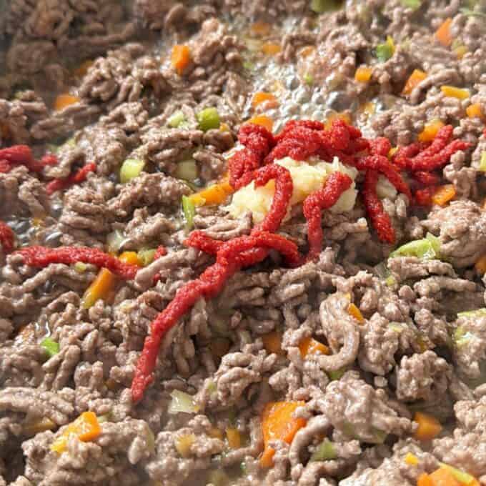 Cheesy Taco Mince and Macaroni Bake Ground beef cooking with chopped carrots and celery, topped with a dollop of minced garlic and lines of tomato paste.