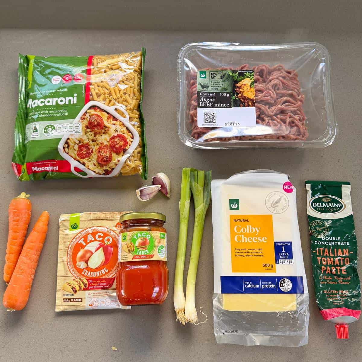 Cheesy Taco Mince and Macaroni Bake Various ingredients on a countertop, including macaroni, beef mince, carrots, garlic, green onions, taco seasoning, pasta sauce, Colby cheese, and tomato paste.