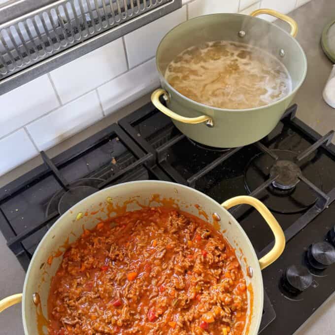 Cheesy Taco Mince and Macaroni Bake A pot of boiling pasta sits next to a pan of simmering meat and tomato sauce on a kitchen stove.