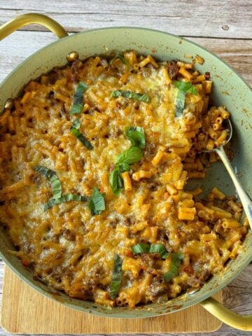 Large green pan filled with baked macaroni and cheese topped with browned cheese and fresh basil, with a spoon resting inside; set on a wooden surface.