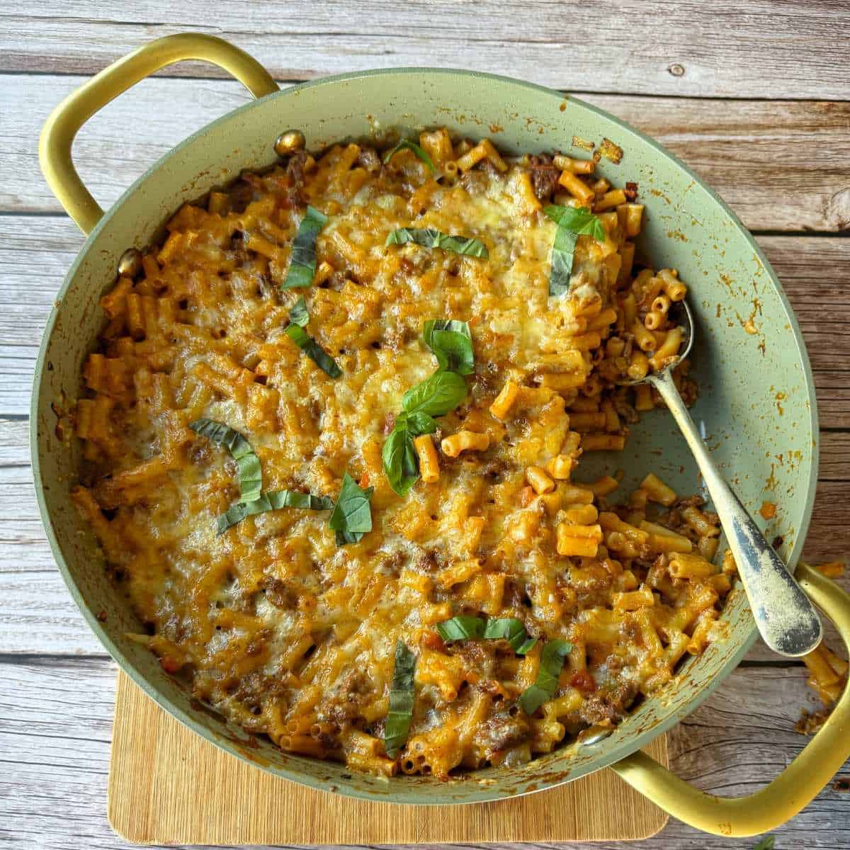 Cheesy Taco Mince and Macaroni Bake Large green pan filled with baked macaroni and cheese topped with browned cheese and fresh basil, with a spoon resting inside; set on a wooden surface.