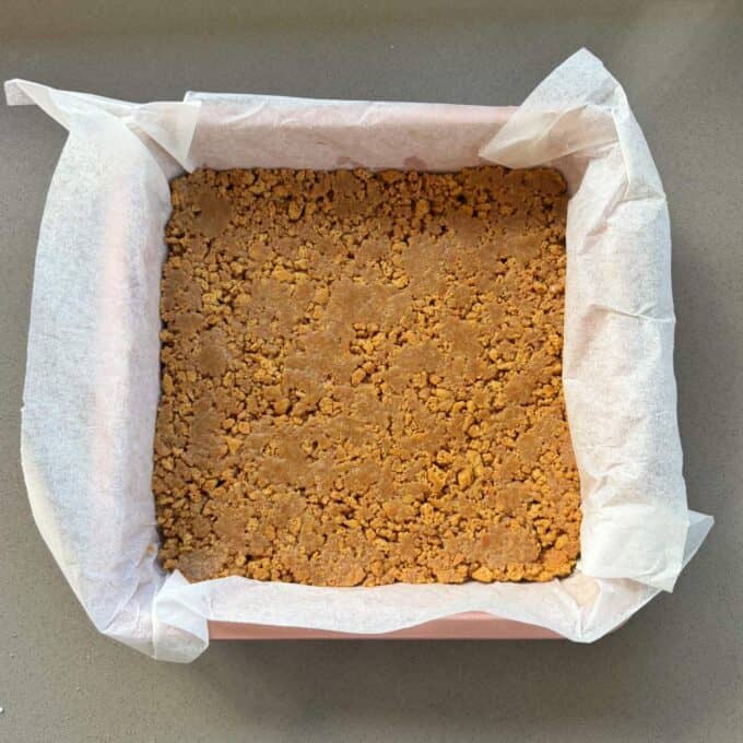 No-Bake Gingernut Slice A square baking pan lined with parchment paper, filled with an even layer of crumbly biscuit crust mixture.