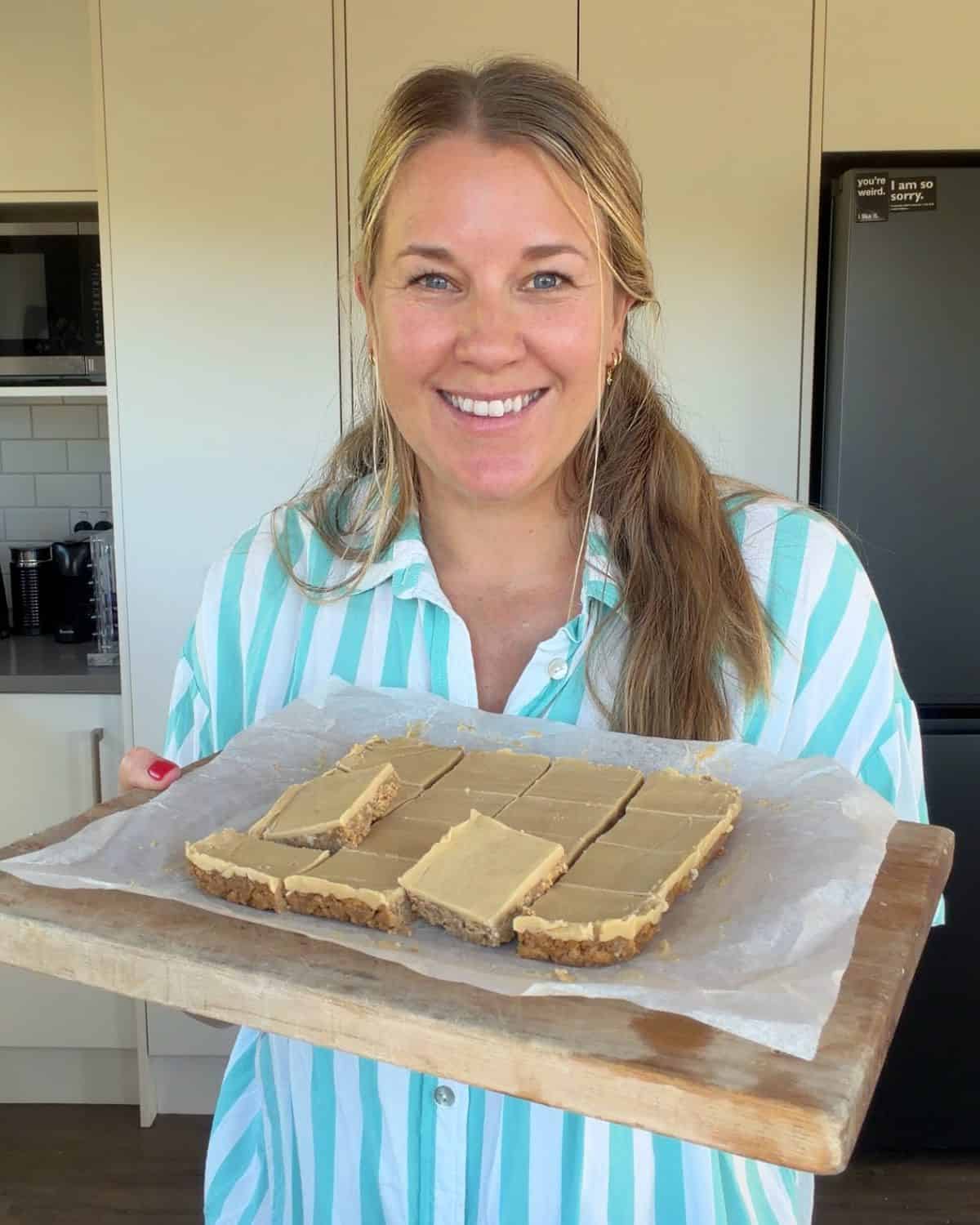 No-Bake Gingernut Slice A woman in a striped shirt holds a wooden tray with neatly cut dessert bars on parchment paper, standing in a kitchen.